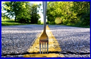 A silver fork stuck tines-down in the yellow stripe in the middle of a tree-shaded road